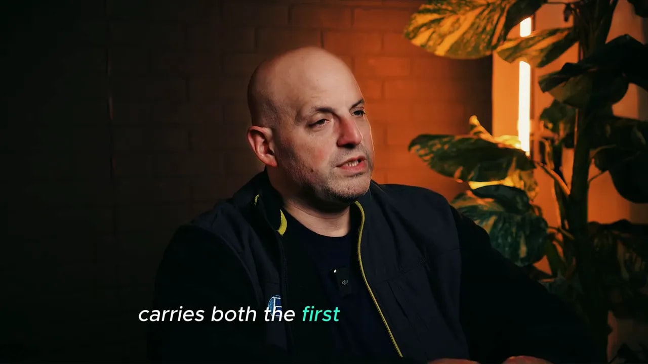 Interview shot showing speaker with on-screen subtitle 'carries both the first' describing how the bank temporarily carries both the first mortgage and interim second loan.